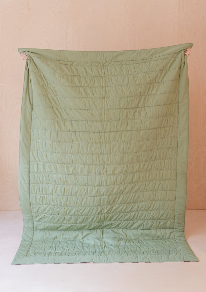 TBCo Celestial Checkerboard quilted cotton throw blanket in sage green, shown from reverse solid side, hung vertically against pink wall
