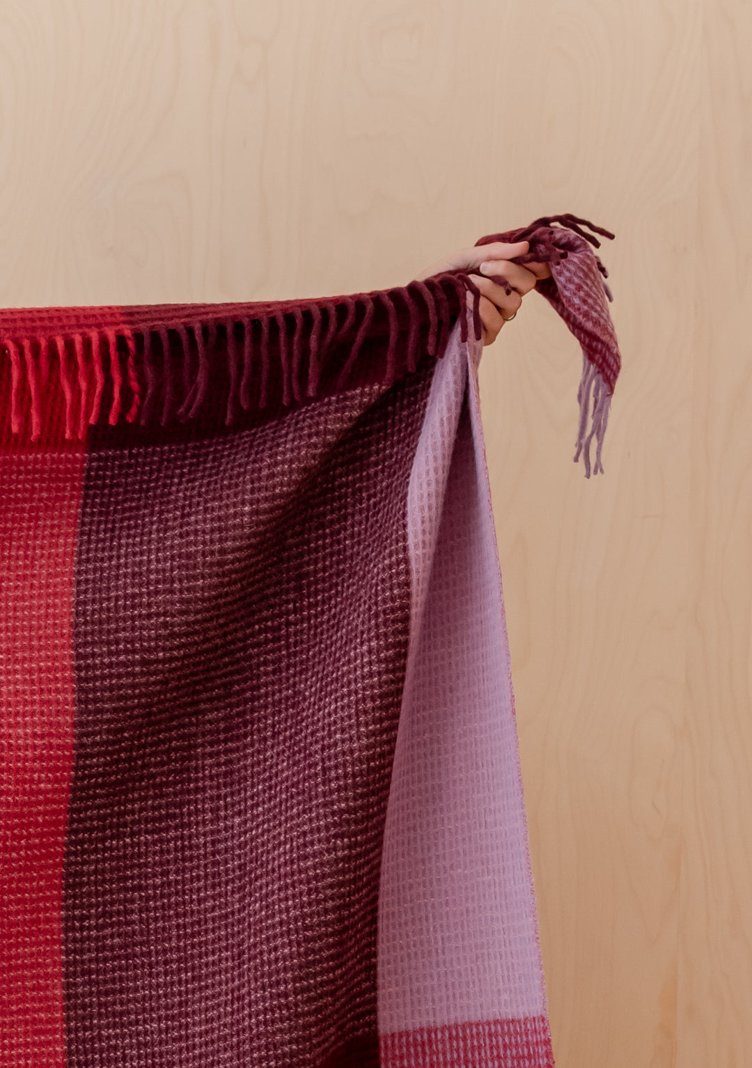 Hand holding TBCo Recycled Wool Blanket in Burgundy Waffle Block Check with fringed edge against wooden background