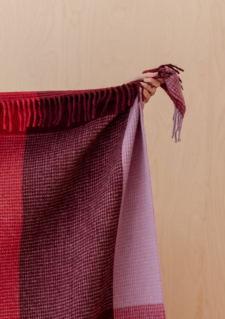 Hand holding TBCo Recycled Wool Blanket in Burgundy Waffle Block Check with fringed edge against wooden background