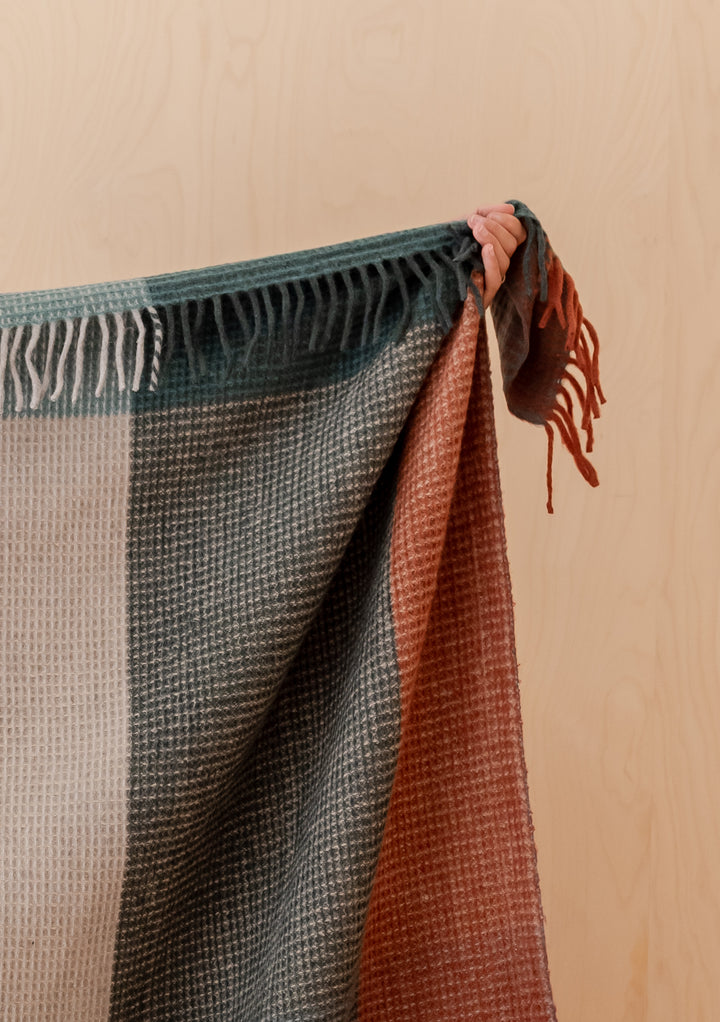 Hand holding TBCo Recycled Wool Blanket in Green Waffle Block Check with fringed edges against wooden background