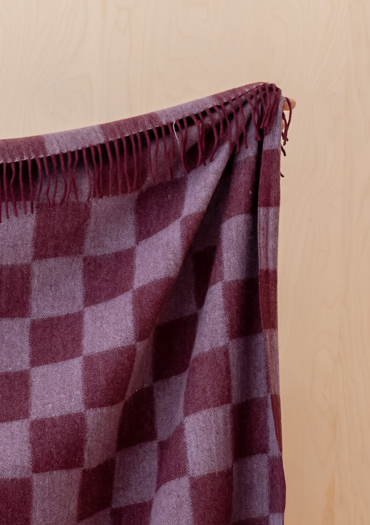 Soft fringed Recycled Wool Blanket in Burgundy & Lilac Checkerboard by TBCo draped against a wooden background