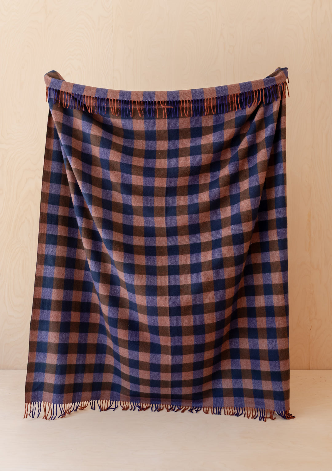TBCo Recycled Wool Blanket in Midnight Gingham with fringed edges hanging against a light wooden background