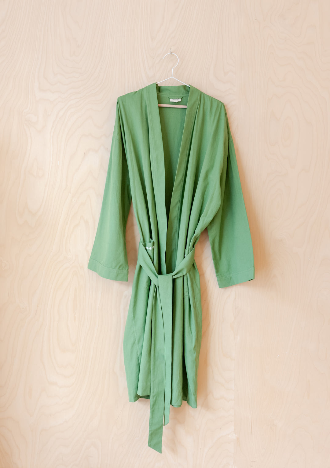 TBCo Cotton & Linen Robe in sage green with integrated belt, hanging on wooden wall, showcasing relaxed fit and flowing fabric