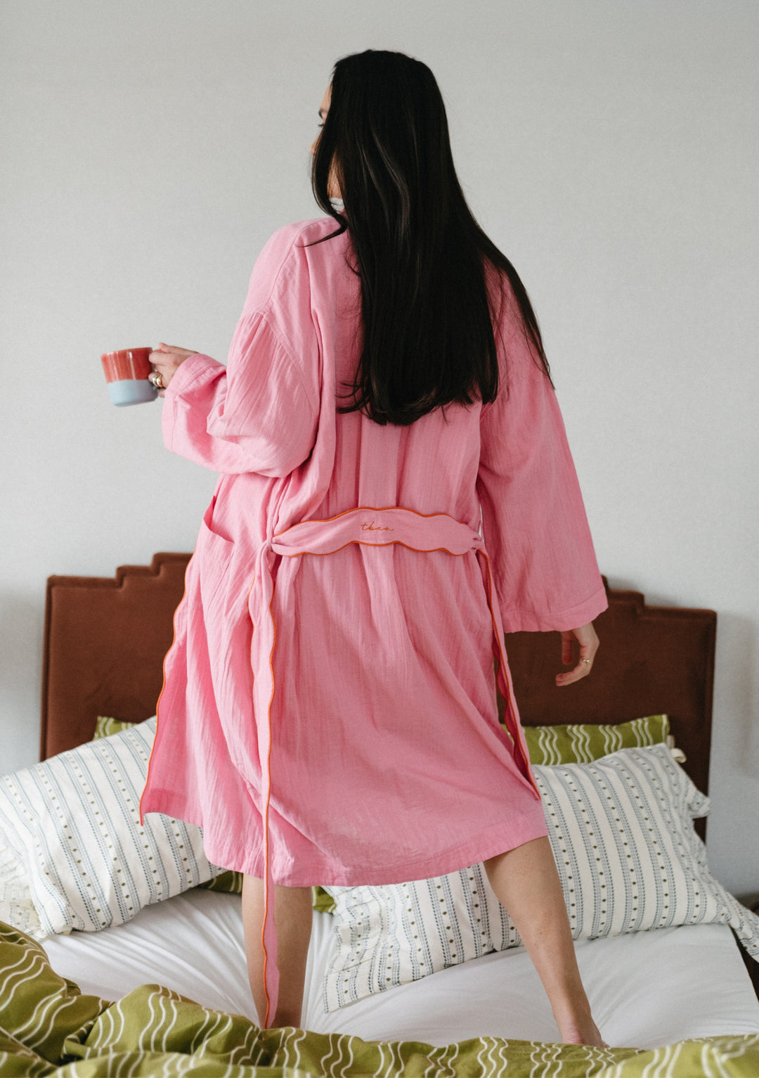 Person wearing TBCo Cotton Robe in Pink Scallop, shown from behind holding a cup while standing on a bed with patterned bedding