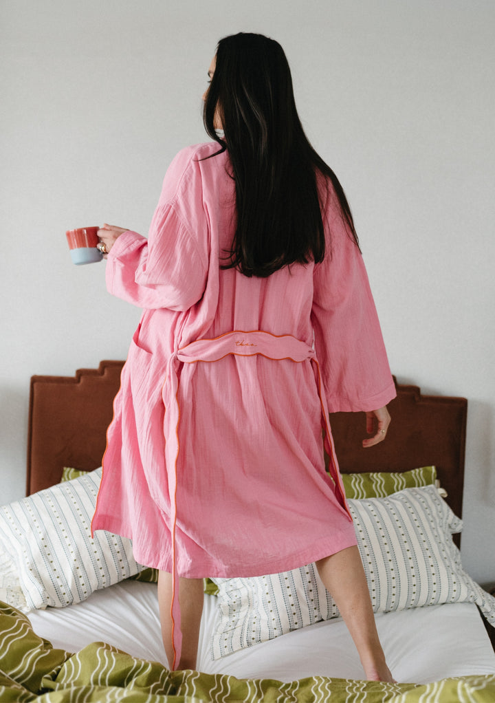Person wearing TBCo Cotton Robe in Pink Scallop, shown from behind holding a cup while standing on a bed with patterned bedding