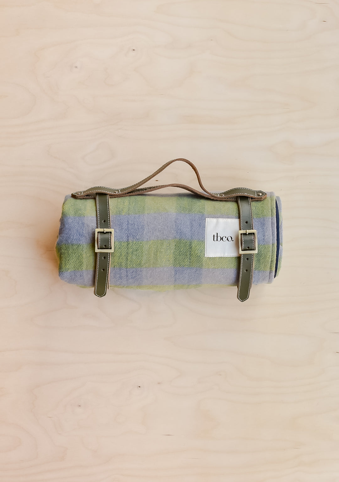 A TBCo recycled wool picnic blanket in green gingham, rolled up and held in a leather TBCo picnic carrier.