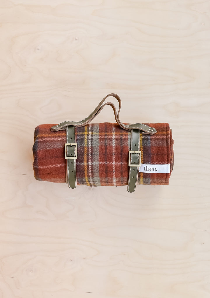 A TBCo recycled wool picnic blanket in Stewart Royal Antique Tartan, rolled up and held in a leather TBCo picnic carrier.