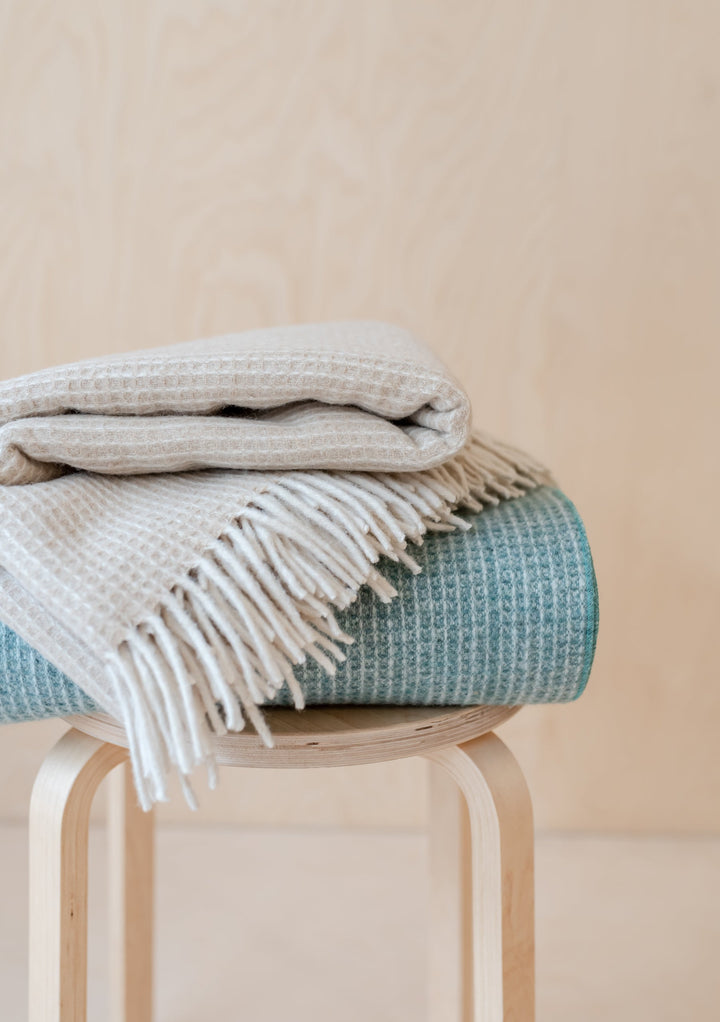 TBCo Recycled Wool Waffle Blanket in Sage, folded neatly on wooden stool showing textured weave and fringed edge detail