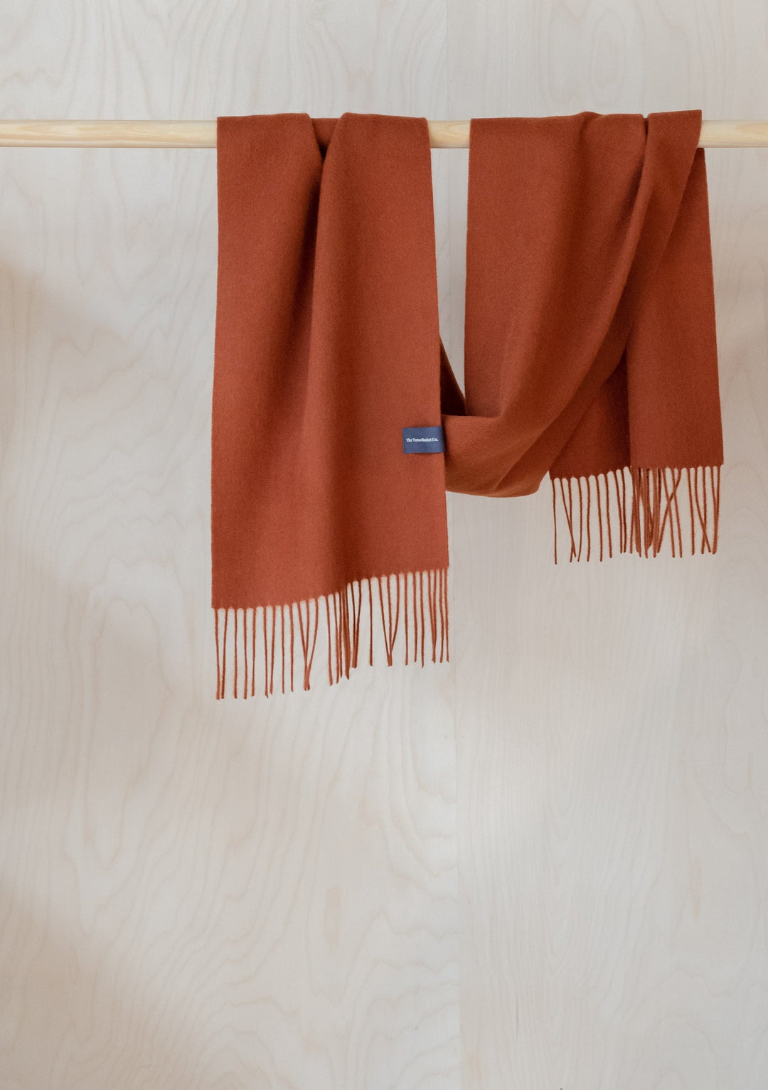 TBCo Lambswool Scarf in Rust Melange draped over wooden rail, showcasing soft texture and fringed ends in warm terracotta colour