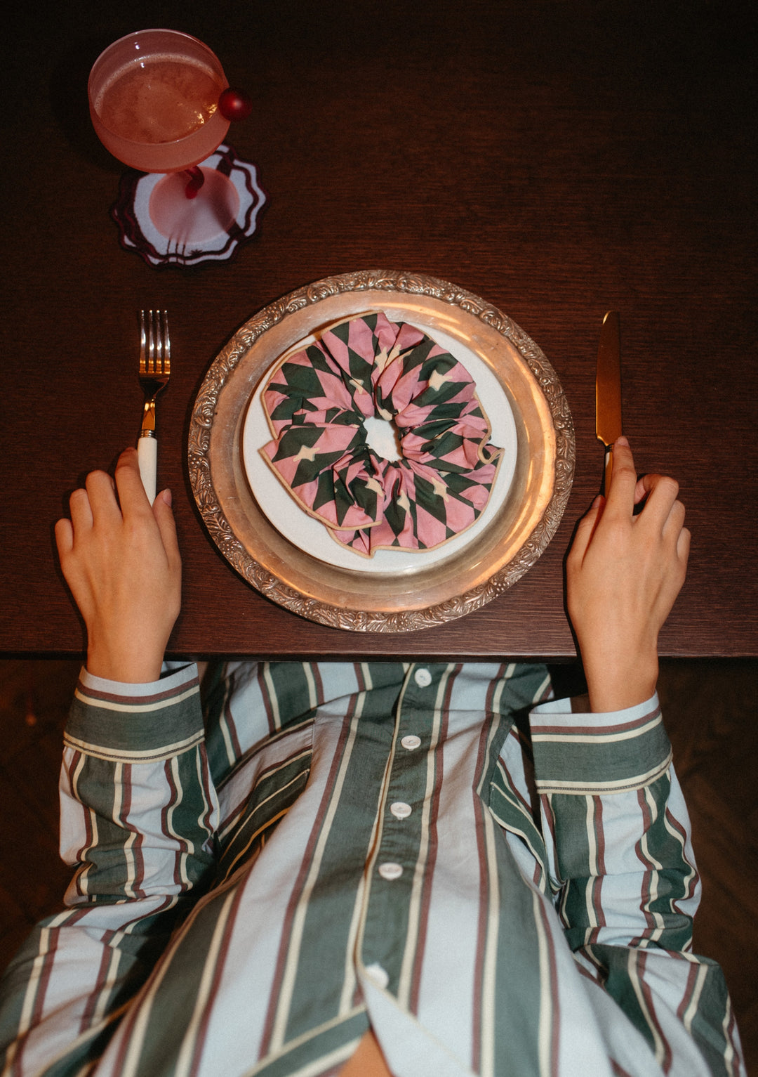 Cotton Oversized Scrunchie in Pink Harlequin by TBCo on a plate with hands holding cutlery and a cocktail glass nearby