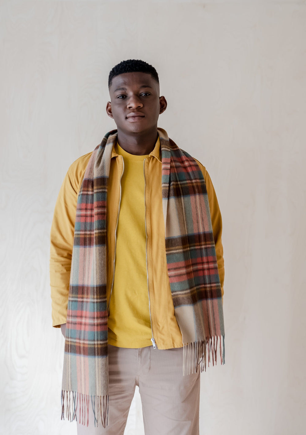A person wearing TBCo's Stewart Dress Antique Tartan Lambswool Scarf with fringed ends, styled with a yellow jacket against light background