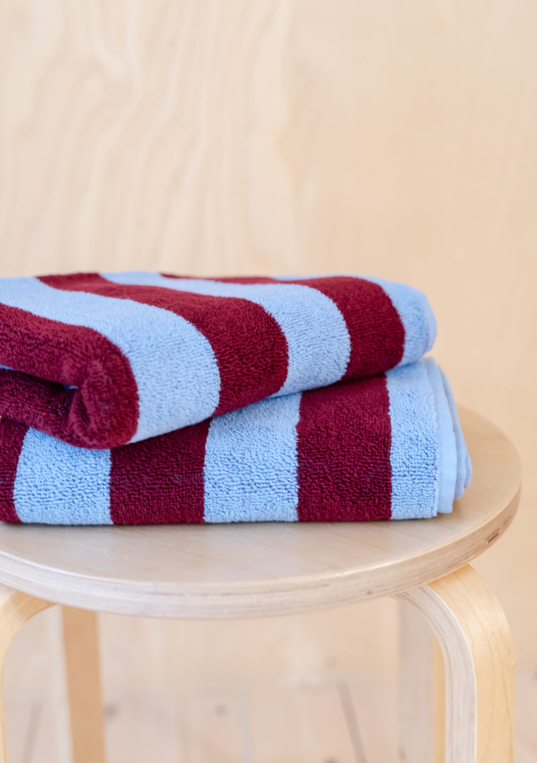 Luxurious TBCo Cotton Towels in Blue Stripe with burgundy stripes, neatly folded on wooden stool showing plush texture