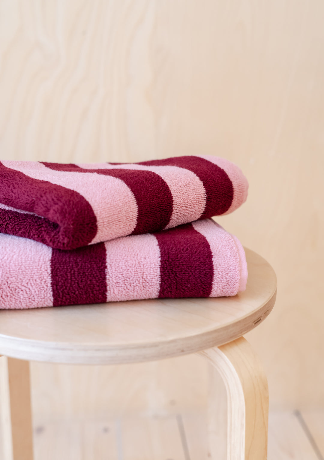 Luxurious TBCo Cotton Towels in Pink Stripe, folded neatly on wooden stool, featuring burgundy stripes and plush cotton texture