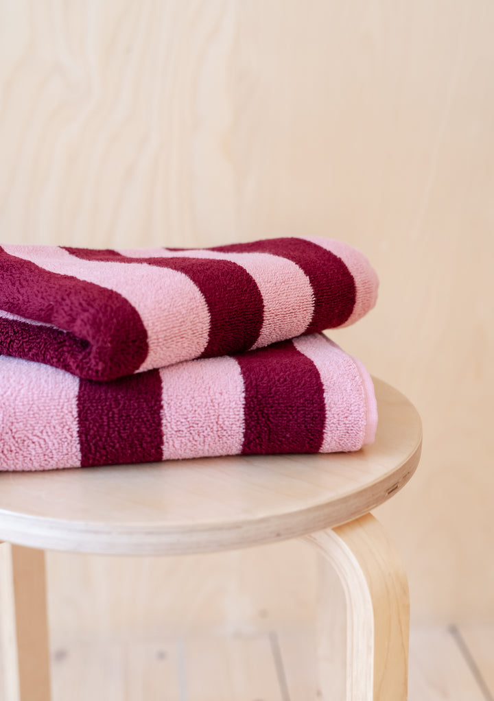 Luxurious TBCo Cotton Towels in Pink Stripe, folded neatly on wooden stool, featuring burgundy stripes and plush cotton texture