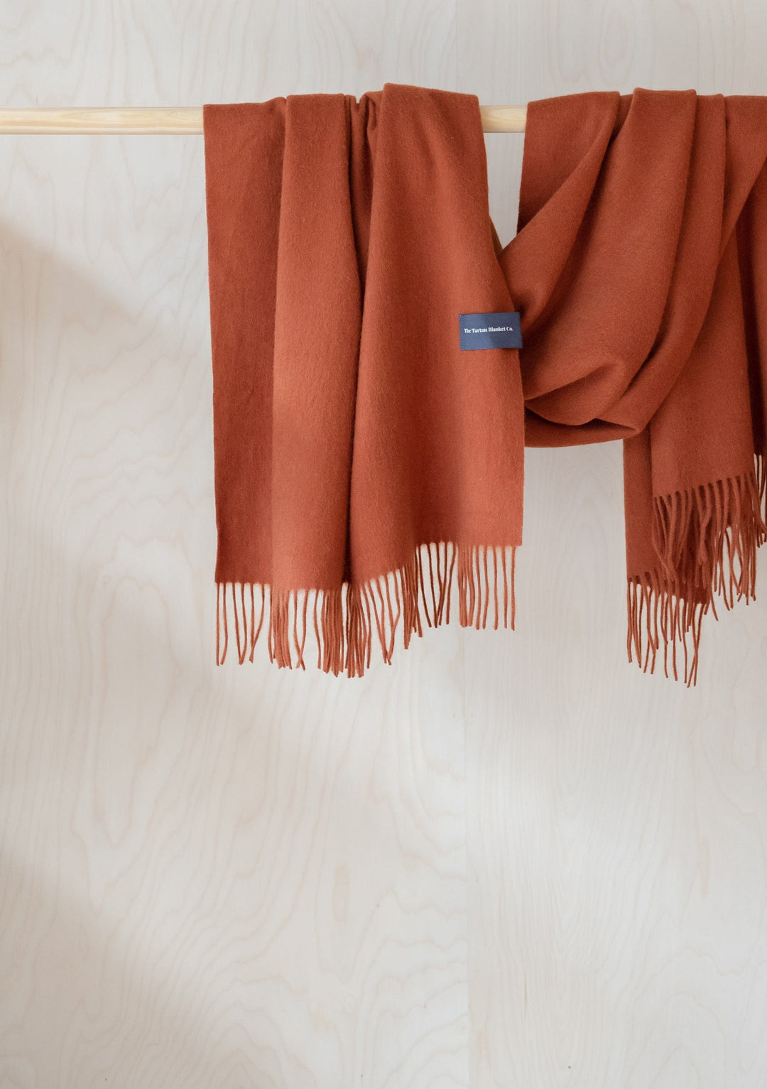 Soft TBCo lambswool blanket scarf in rust melange with fringed edges, draped elegantly over wooden rail against white backdrop
