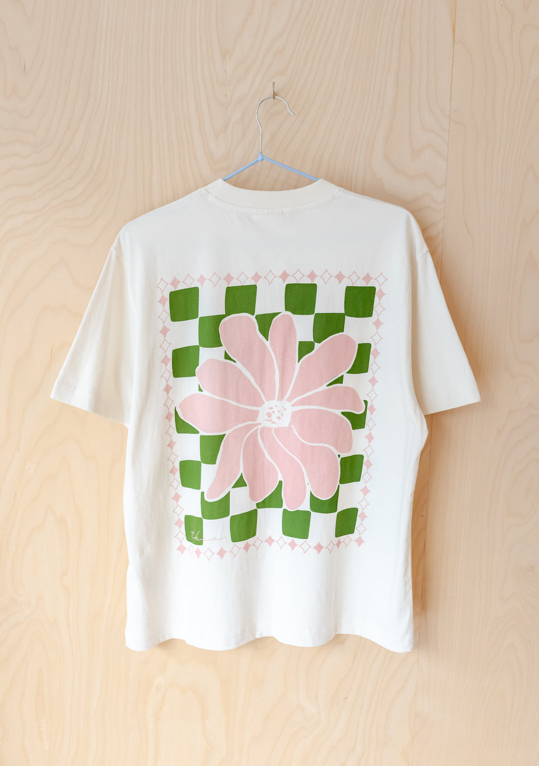 TBCo Cotton T-Shirt in Ecru Flora featuring pink flower on green checkerboard pattern, hanging on wooden wall background