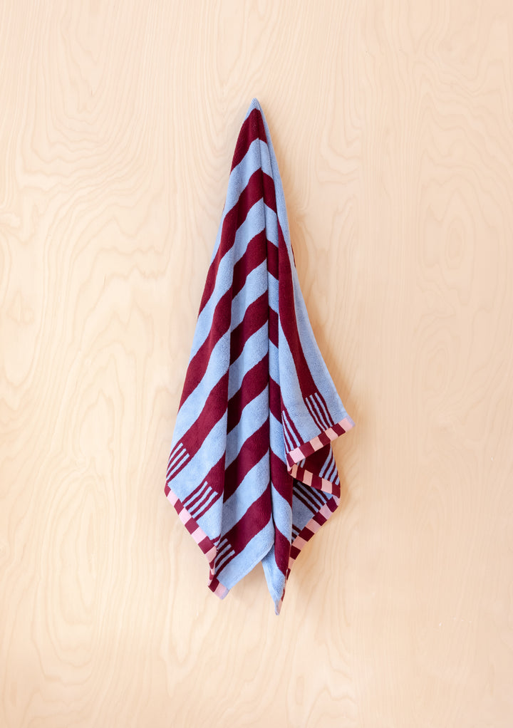 TBCo Cotton Towels in Blue Stripe with burgundy diagonal pattern and checkered border, hanging elegantly against beige surface