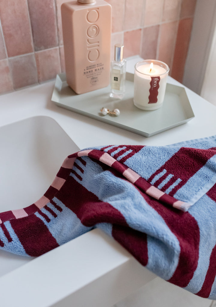TBCo Cotton Towels in Blue Stripe with burgundy pattern, displayed on white bathtub edge alongside luxury bath products and candle