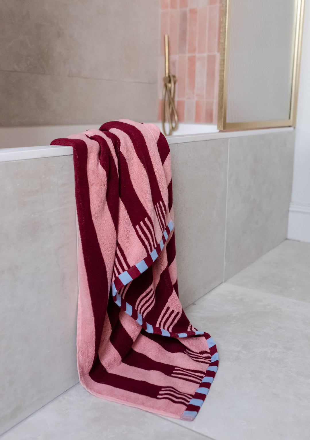 TBCo Cotton Towels in Pink Stripe draped over modern bathroom ledge, featuring burgundy stripes and blue checkerboard trim