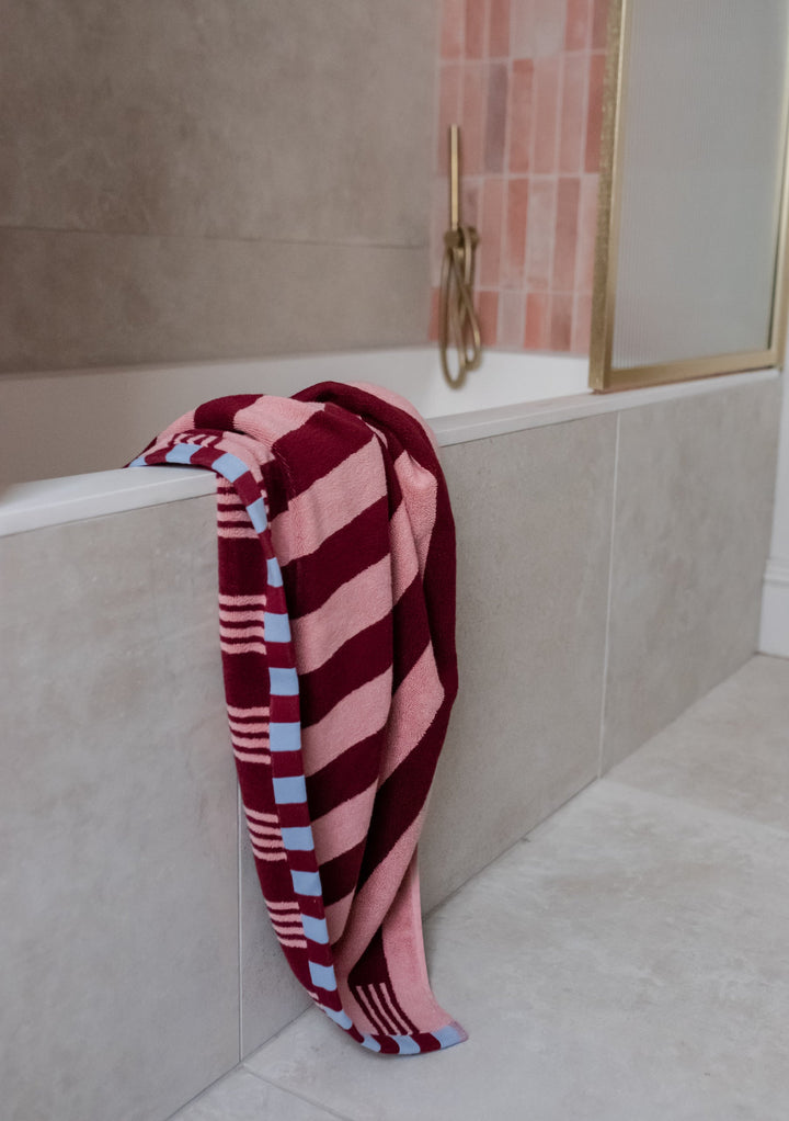 TBCo Cotton Towels in Pink Stripe draped over modern bathroom ledge, featuring burgundy stripes and blue checkerboard trim