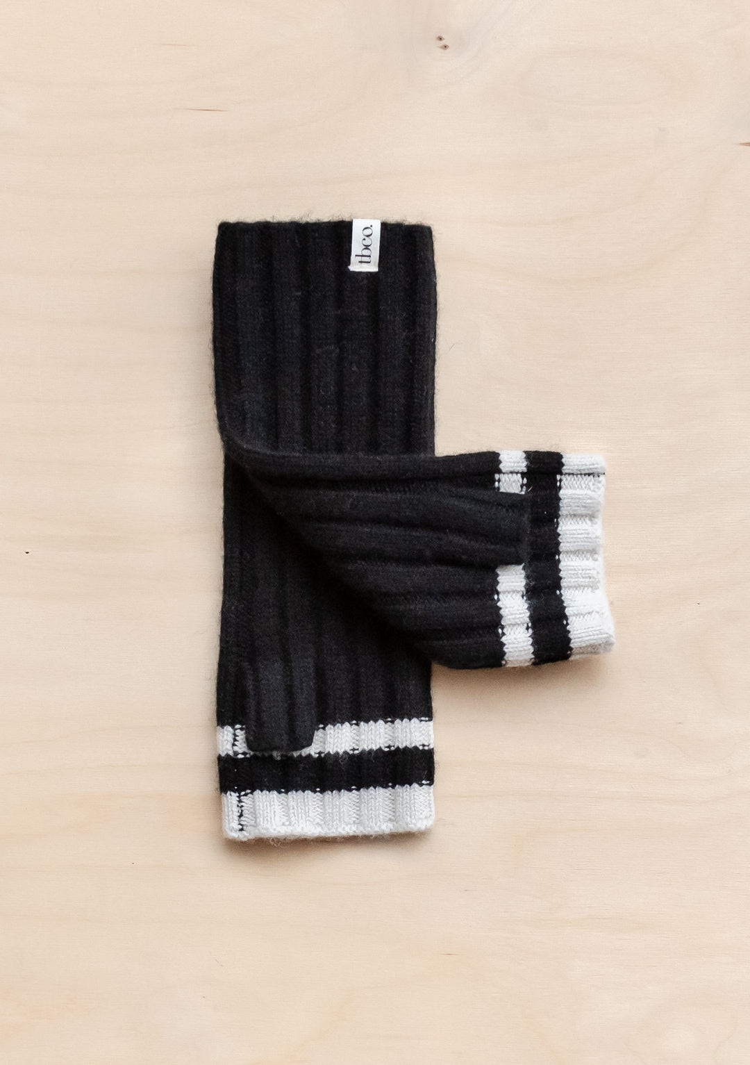 TBCo Cashmere & Merino Wrist Warmers in black ribbed knit with cream striped trim, displayed flat on beige surface