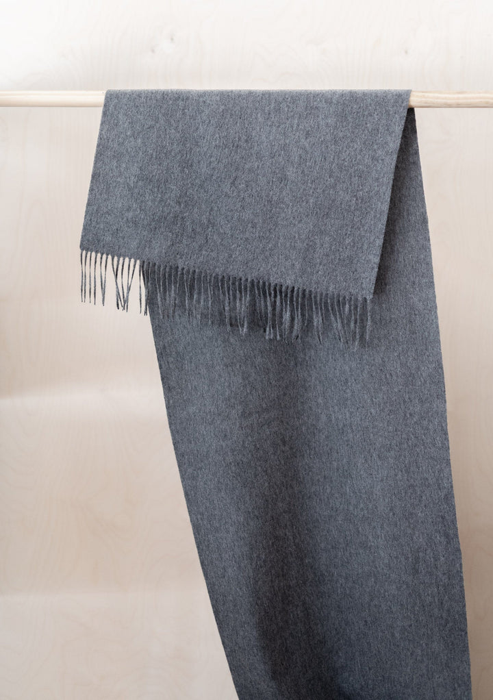 Luxurious TBCo Lambswool Oversized Scarf in Charcoal Melange draped over wooden rail showing fringed detail and soft texture