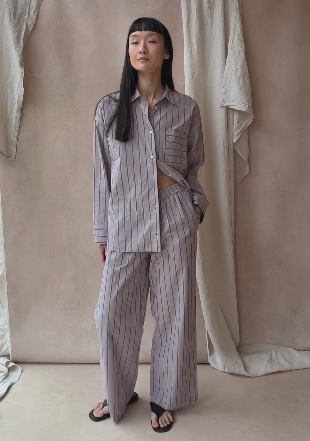 A model poses against a neutral ply backdrop, wearing TBCo matching lilac stripe shirt and trousers.