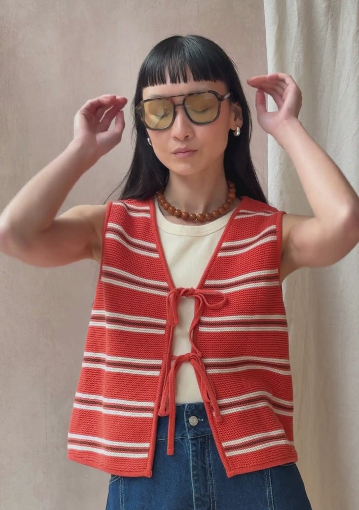 A model poses against a neutral ply backdrop, wearing TBCo orange stripe vest with bow ties on the front, a cream vest, jeans and orange bead necklace.