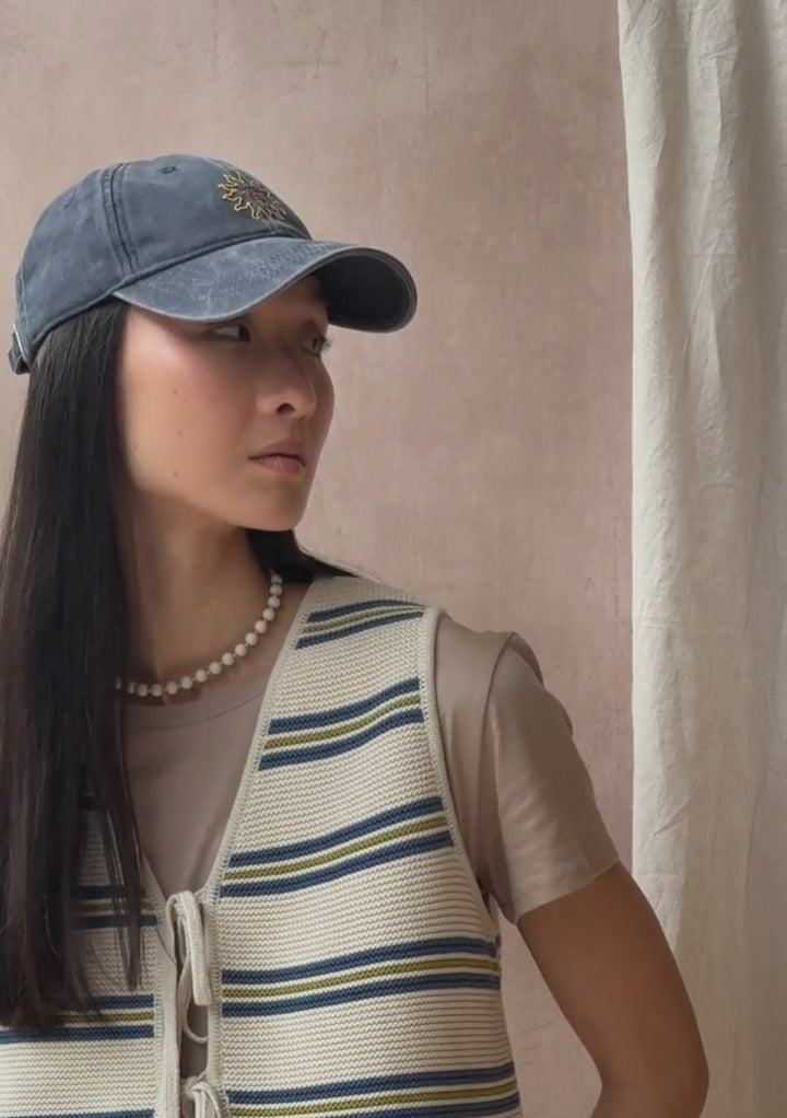 A model poses against a neutral ply backdrop, wearing a TBCo cream and blue stripe vest and blue baseball cap.