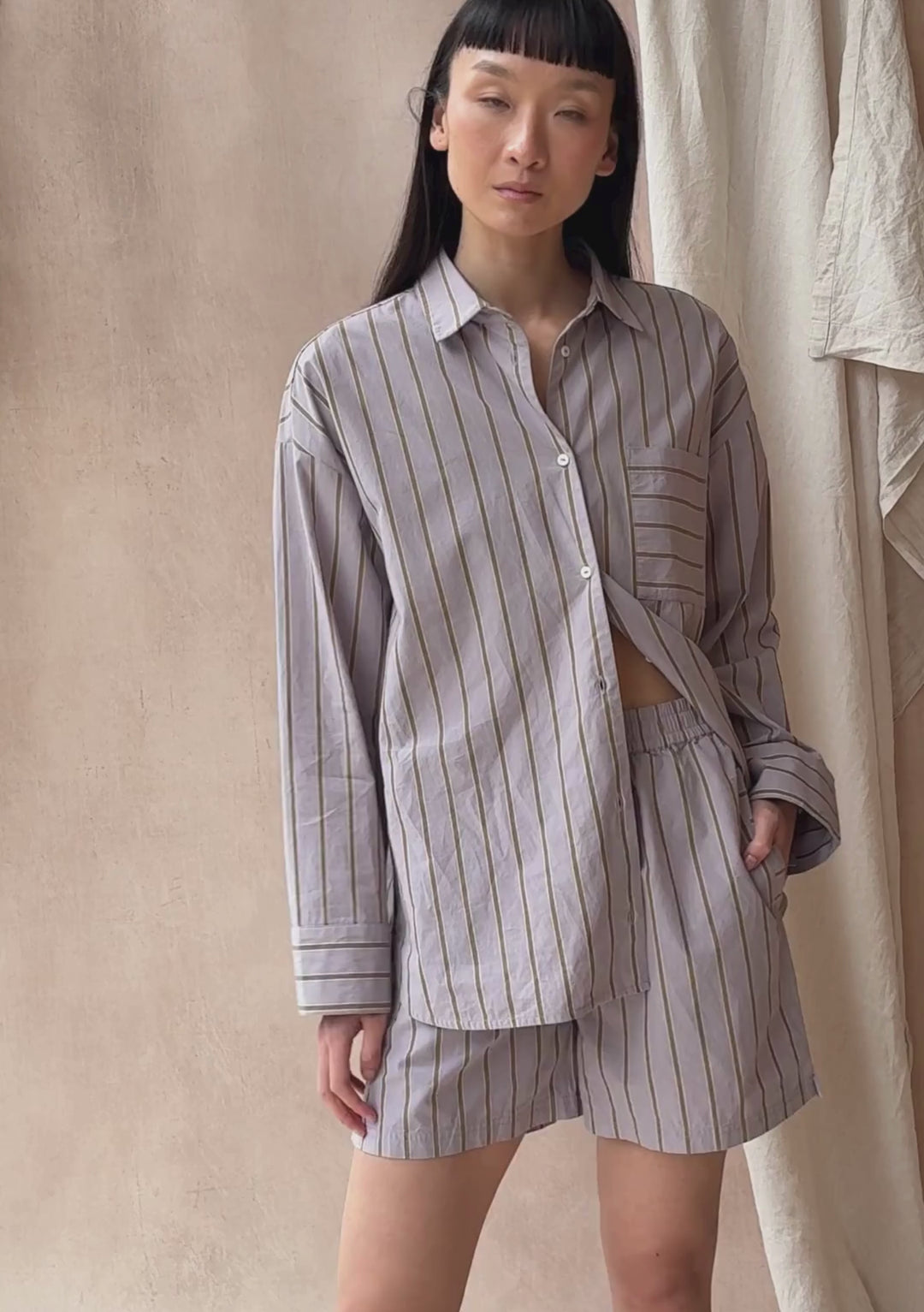 A model poses against a neutral ply backdrop, wearing TBCo matching lilac stripe shirt and shorts.