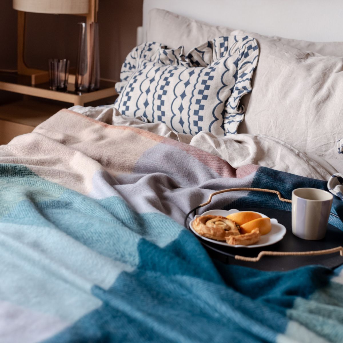TBCo Recycled Wool Apricot Framed Check blanket in a check pattern draped over a cosy bed with TBCo Cotton textured Blue ruffle cushions. A perfect setting for relaxed mornings with coffee and pastries.