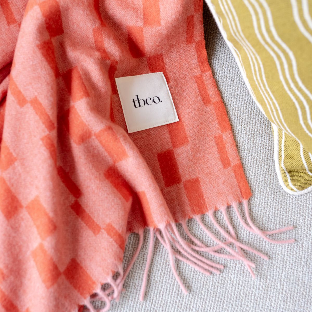 Close-up of the TBCo label on a Lambswool Tangerine Orange Checker Stripe Blanket with a Cotton Green Wave Cushion beside it. 
