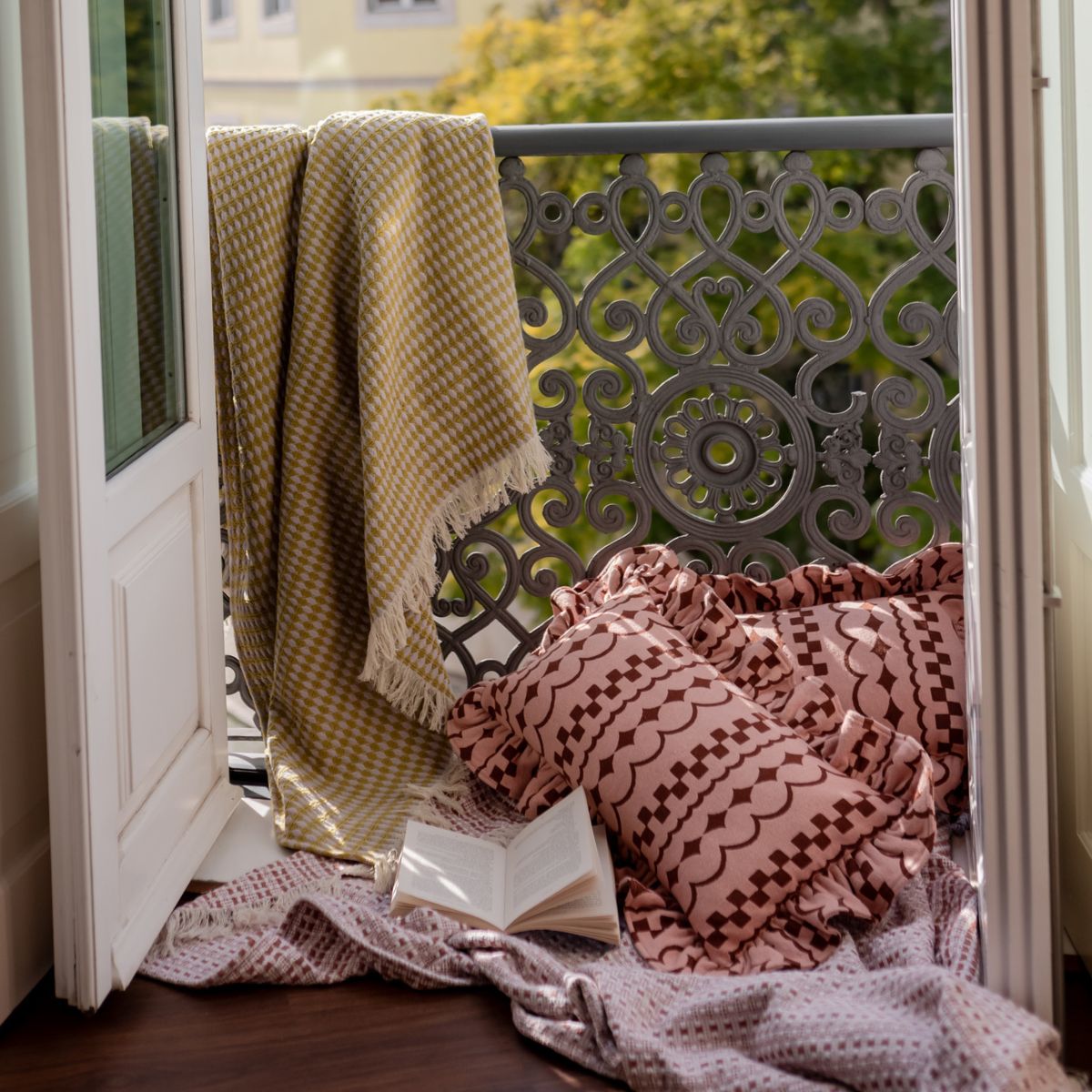 TBCo Cotton Olive Argyle Throw draped over a balcony rail with textured TBCo cotton pink ruffle cushions and Cotton Pink Geometric Throw under it on the floor. A perfect setting for relaxed mornings reading a book. 