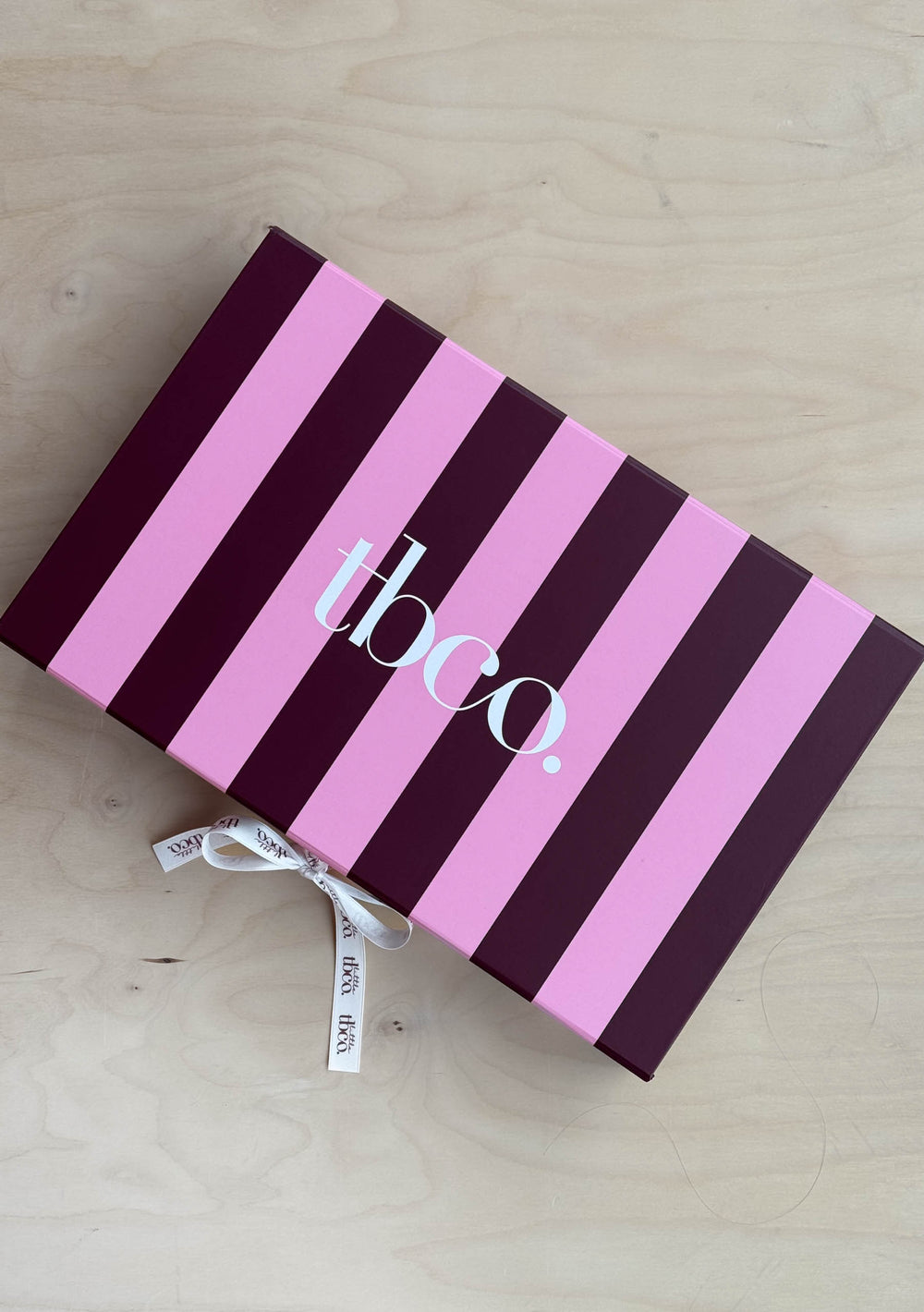 Gift Box For Her by TBCo striped in pink and burgundy with branded ribbon on natural wooden surface