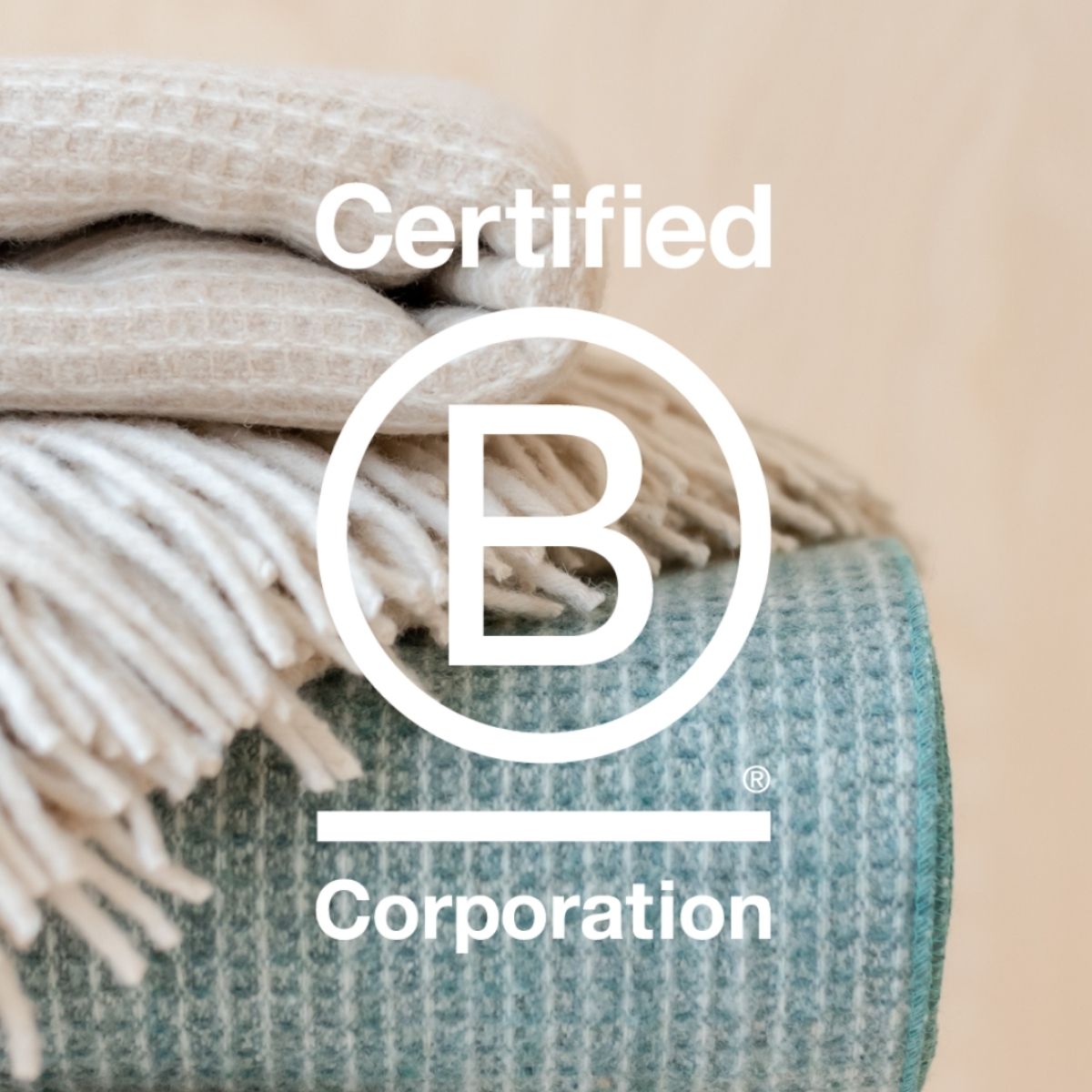 TBCo. Certified B Corporation logo overlaid on a folded Recycled Wool Waffle Blanket stack in soft cream and teal tones. A symbol of ethical craftsmanship, sustainability, and responsible business practices in textile production.