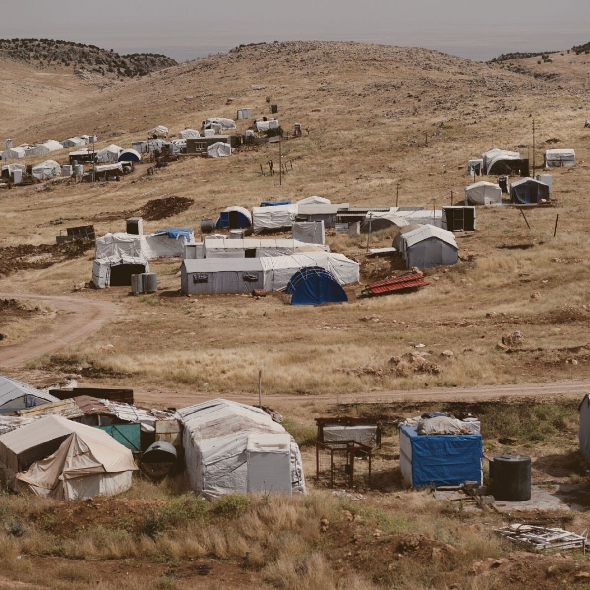 TBCo Impact Pledge donating to those in need. Refugee camp in a remote, arid landscape with makeshift white and blue shelters scattered across a dry hillside under a hazy sky