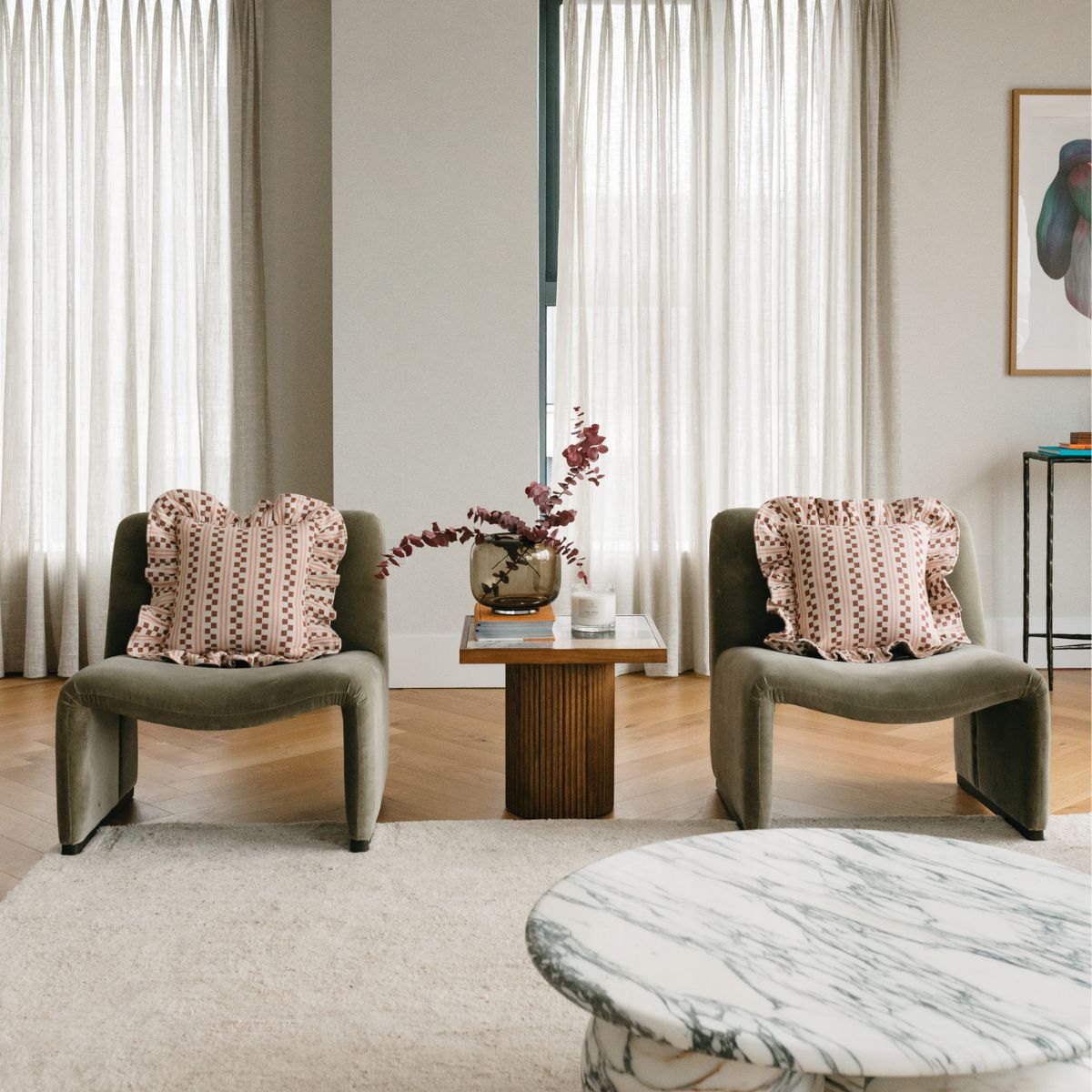 TBCo ruffled cotton cushion covers in a soft pink and red geometric pattern, elegantly styled on olive-green lounge chairs in a modern living room. A chic and cosy interior setting with natural light and minimalist decor.