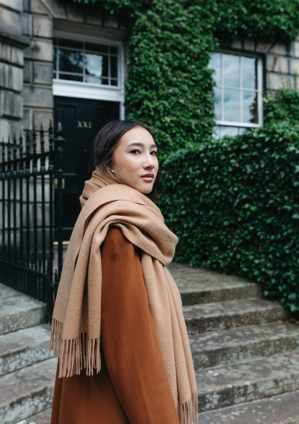 Luxurious TBCo camel cashmere blanket scarf draped elegantly against stone building and ivy backdrop in Edinburgh