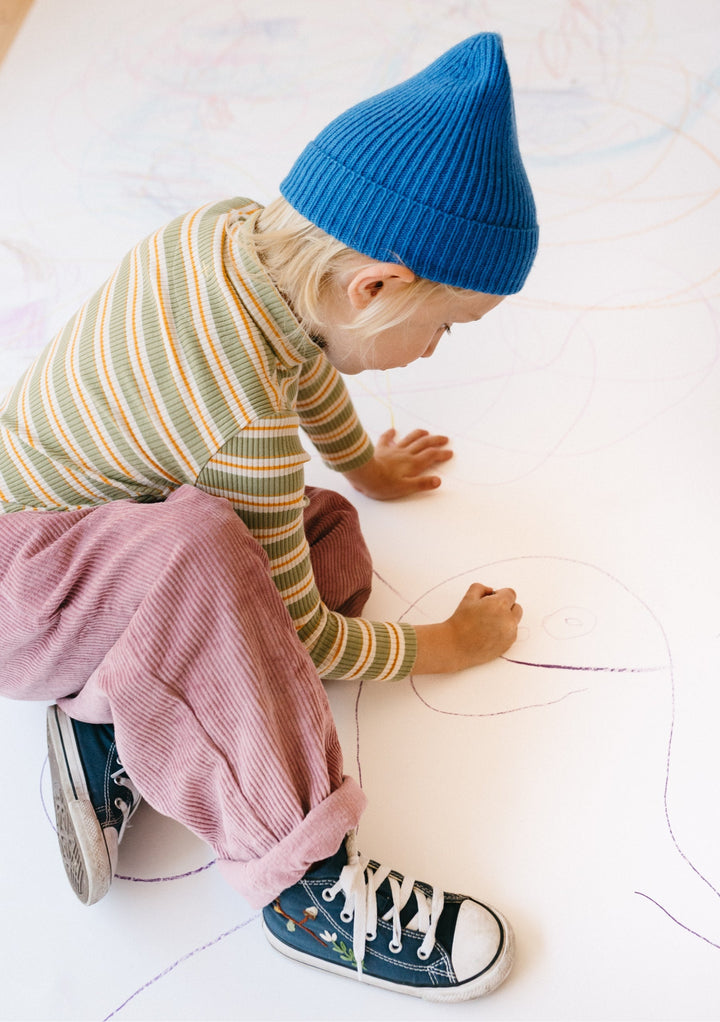 Young child wearing TBCo Merino Wool Kids Beanie in Blue while drawing on the floor, showing the beanie's cosy fit