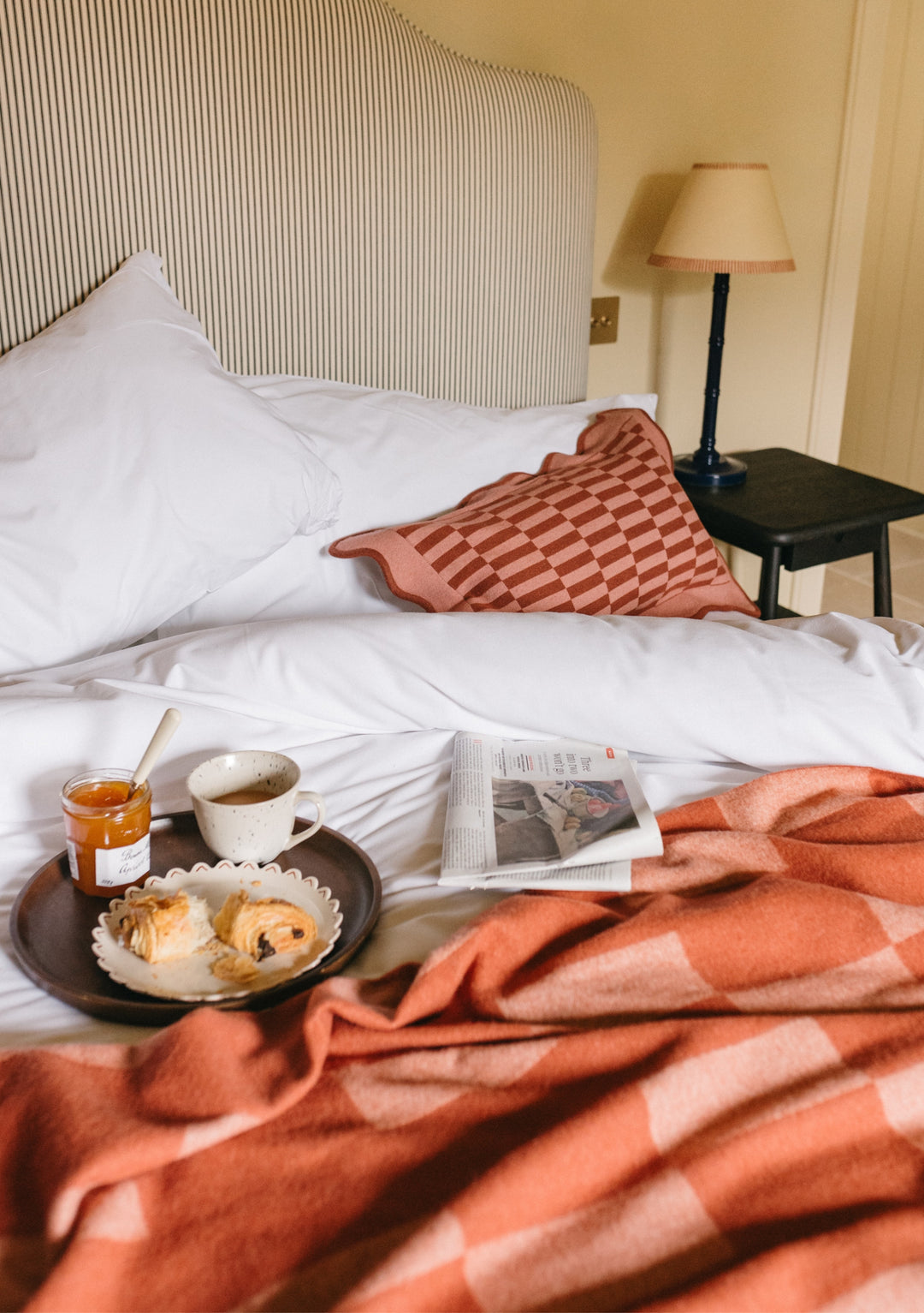 Cosy bedroom scene featuring TBCo Mix & Match Blanket with Cushion Set in coral, styled with breakfast tray on white bedding