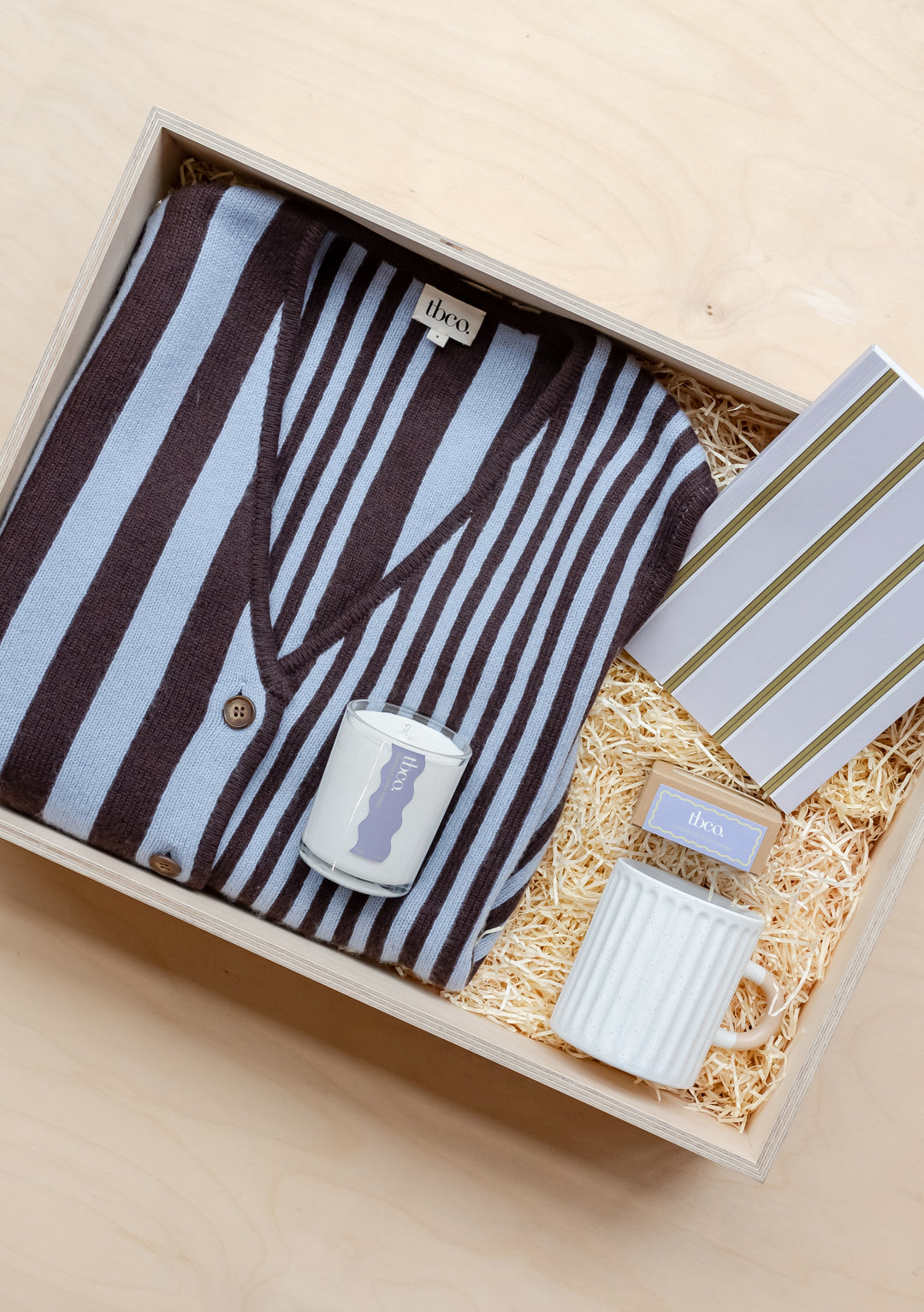 TBCo Ultimate Gift Box with striped pajamas, candle, and ceramic mug nestled in wooden box with straw filling