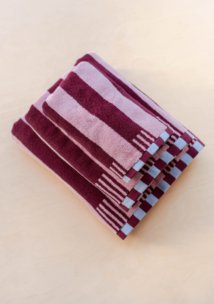 Stack of TBCo Terry Cotton Towels in Burgundy Stripe displaying plush texture and blue checkerboard edge detail on wood surface