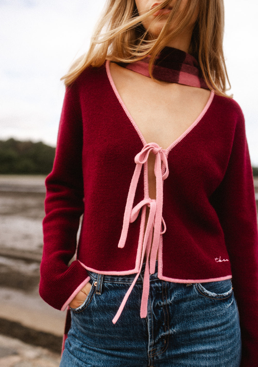 TBCo Merino Wool Tie Cardigan in Burgundy, featuring a V-neck, pink trim, and adjustable ties, styled over jeans with a matching scarf at a riverside setting.
