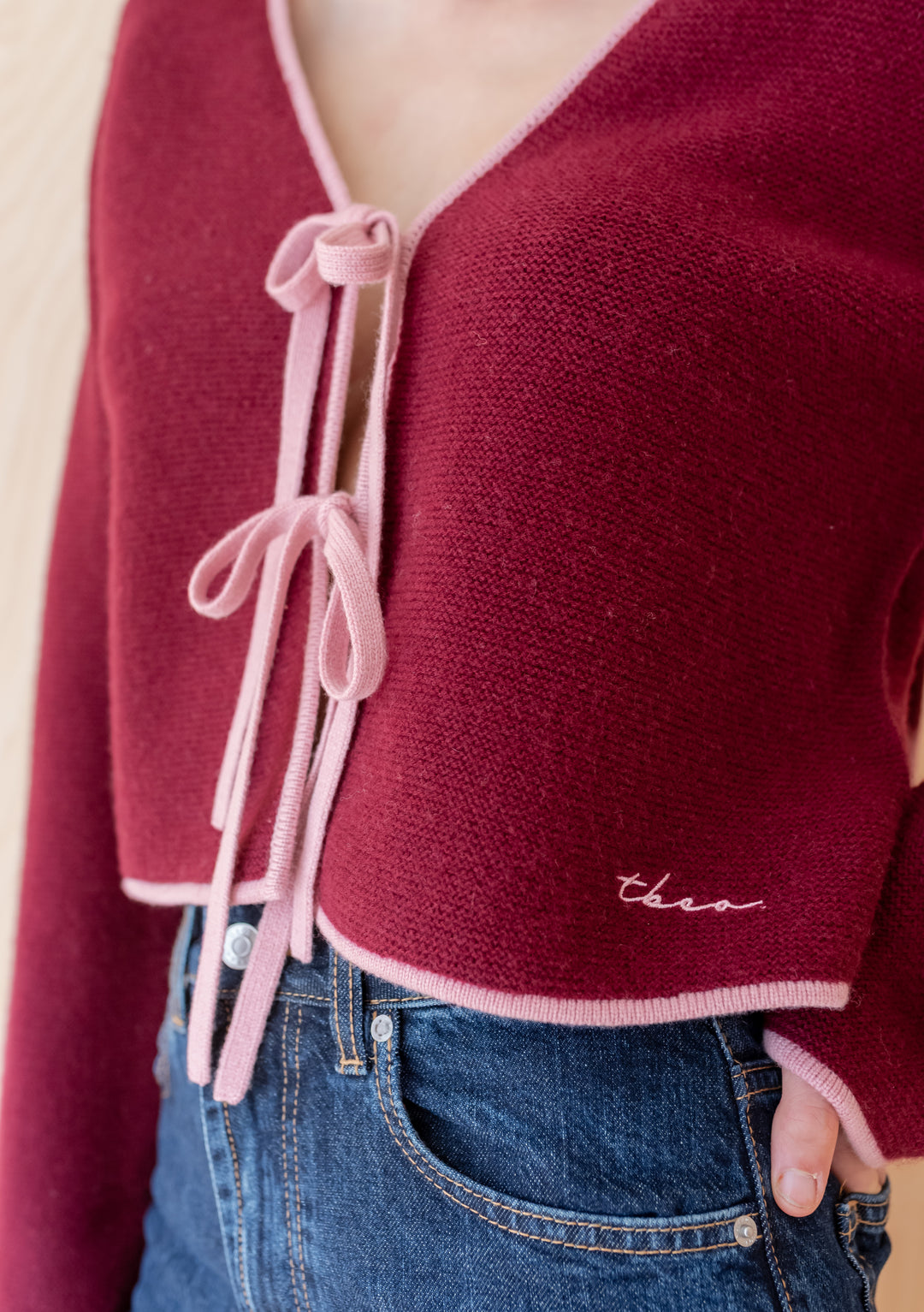 TBCo Merino Wool Tie Cardigan in Burgundy, featuring soft pink trim and tie-strings, worn with medium-wash jeans, showcasing embroidered hem detail.