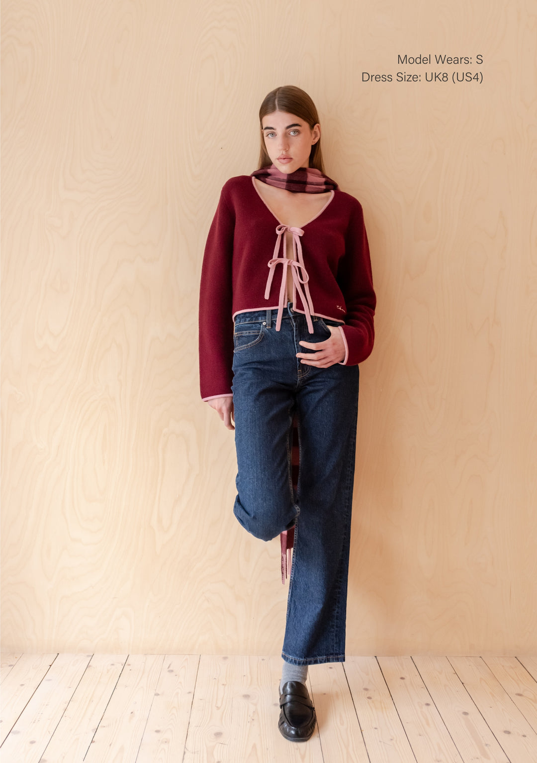 Model wearing TBCo Merino Wool Tie Cardigan in Burgundy with pink trim and ribbon ties, paired with a plaid scarf, blue jeans, grey socks, and black loafers.