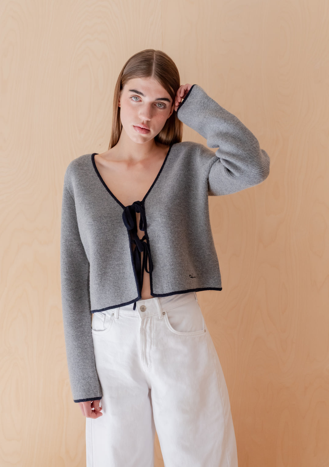 Young woman in TBCo Merino Wool Tie Cardigan in Grey with bow-tie closures, paired with high-waisted white jeans, standing confidently against a light-wood backdrop.