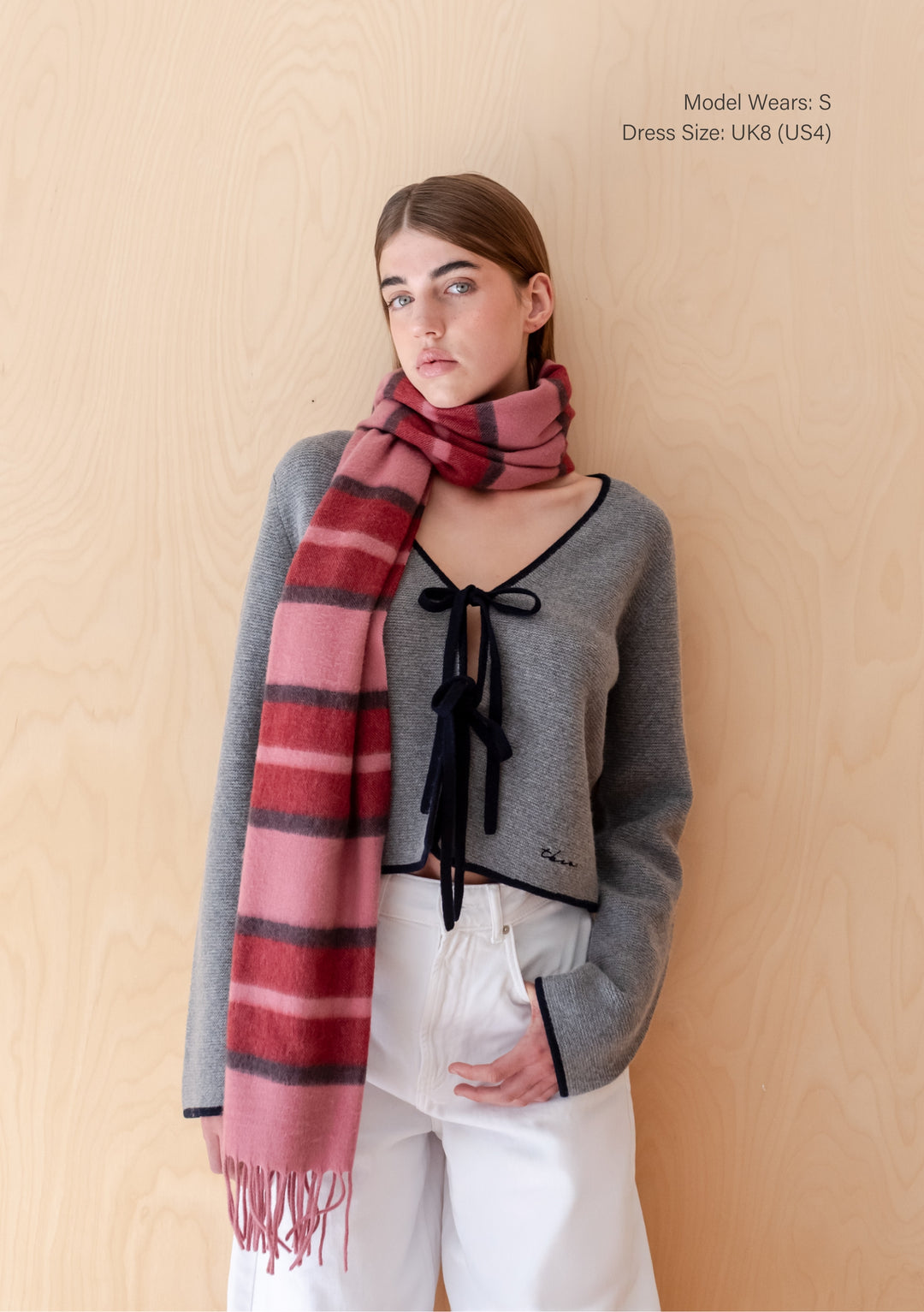 TBCo Model wearing Merino Wool Tie Cardigan in Grey with black-trimmed edges and tie closures, paired with a TBCo pink-red striped scarf and white wide-leg trousers.