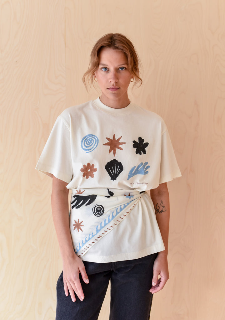 TBCo Chiara Perano Artist Series Cotton Square Triangle Neck Scarf worn as a belt and the Cotton T-shirt on a model standing in front of a wooden background. 