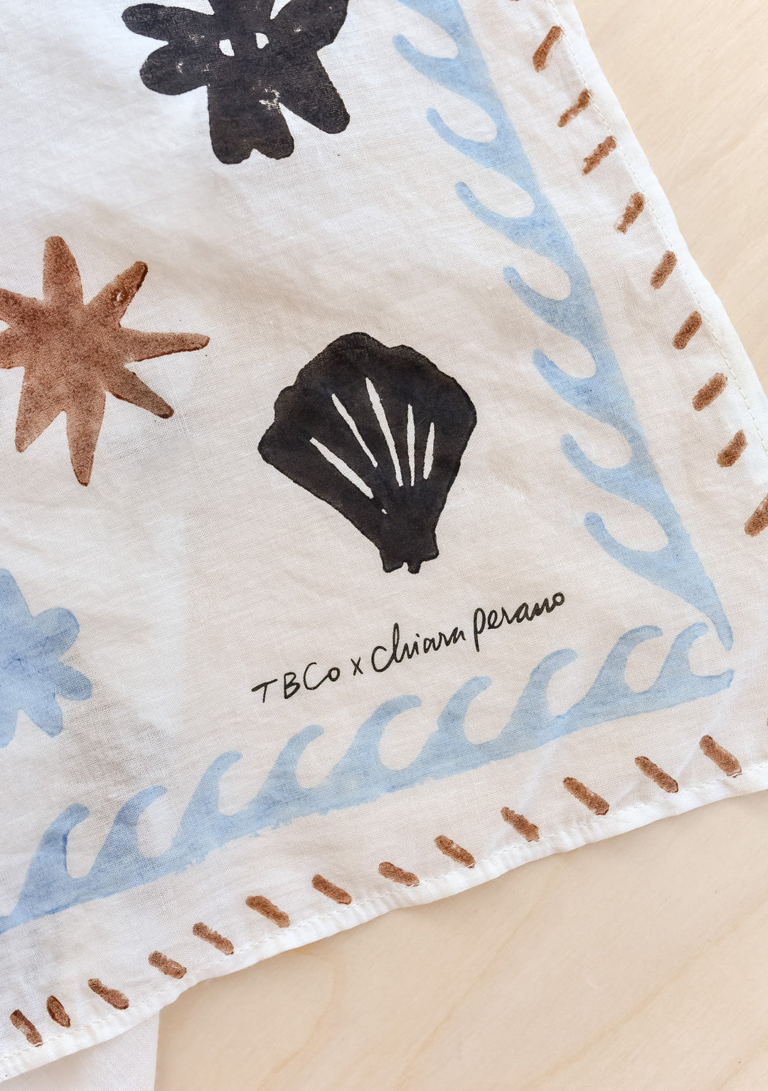Signature corner detail of TBCo Chiara Perano Artist Series Cotton Square Triangle Neck Scarf laid flat on a wooden background