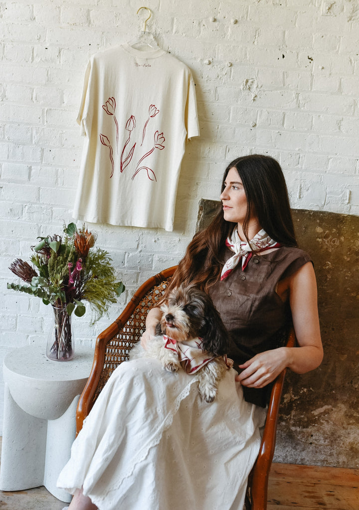 Female artist wearing TBCo x Chiara Perano Artist Series white and red floral cotton neck scarf, seated with a small dog also wearing the matching scarf, in a rustic interior with a floral bouquet on a side table and a cream-colored floral line-art T-shirt hanging on a white brick wall in the background.